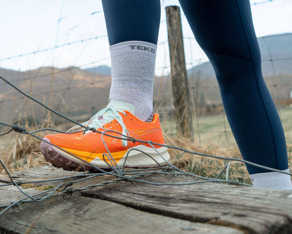 Person wearing blue leggings and orange shoes sitting on a wooden post with a mountainous background.