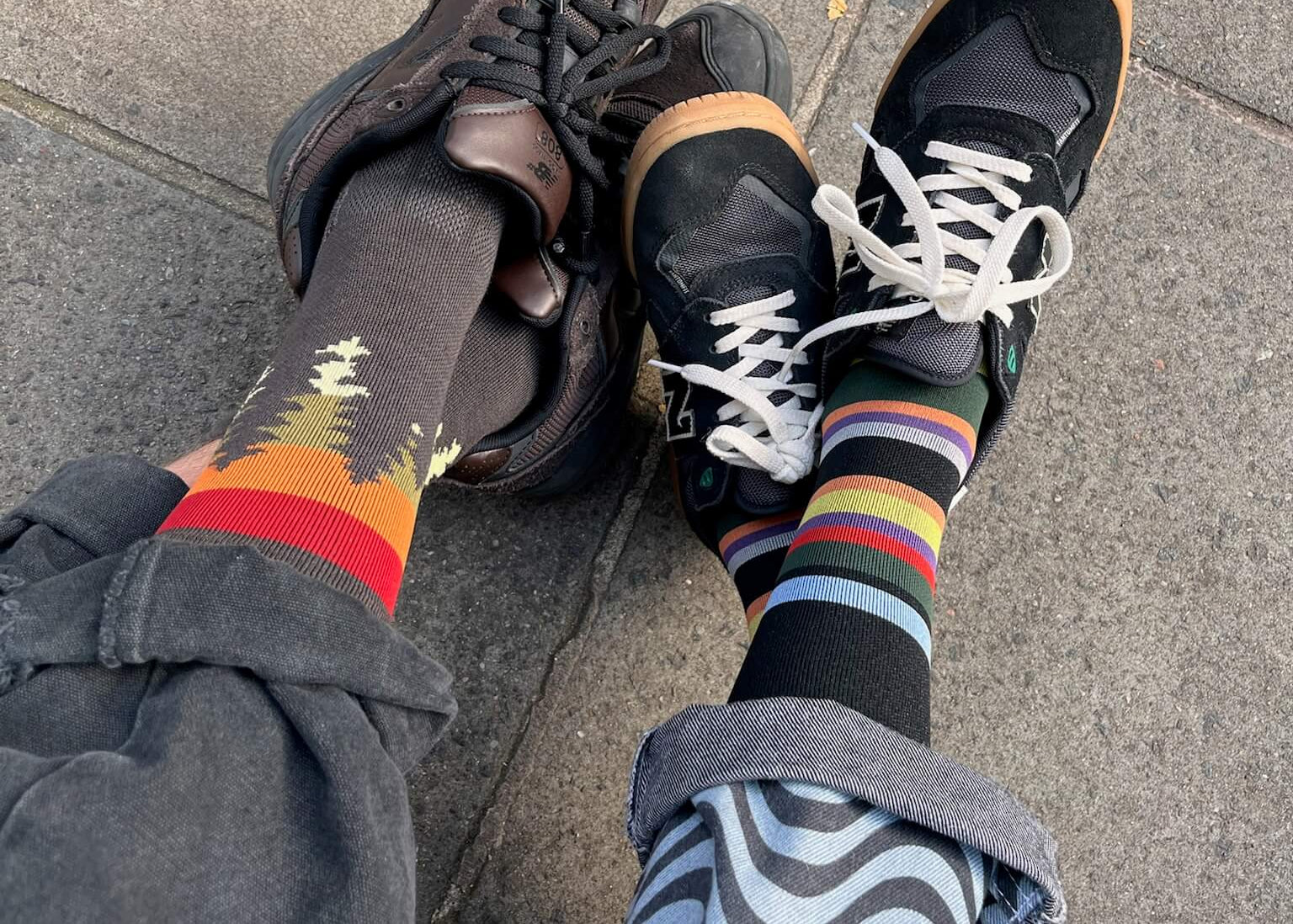 Two pairs of feet wearing colorful socks and black sneakers on a pavement.
