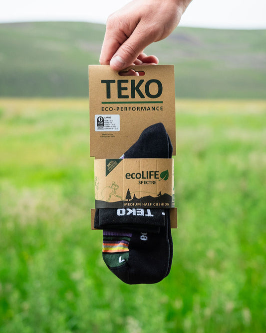 A hand holds a package of TEKO Eco Performance athletic socks against a blurred green field background. The packaging is brown kraft paper with black and green branding, and the socks are black with white branding.