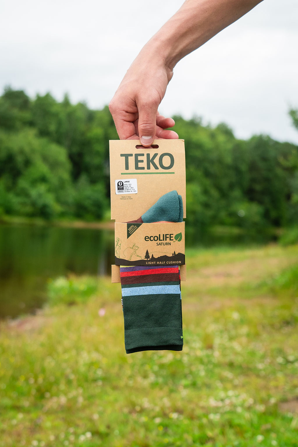 A hand holding a package of TEKO EcoLife outdoor socks against a blurred outdoor background of green trees and grass.