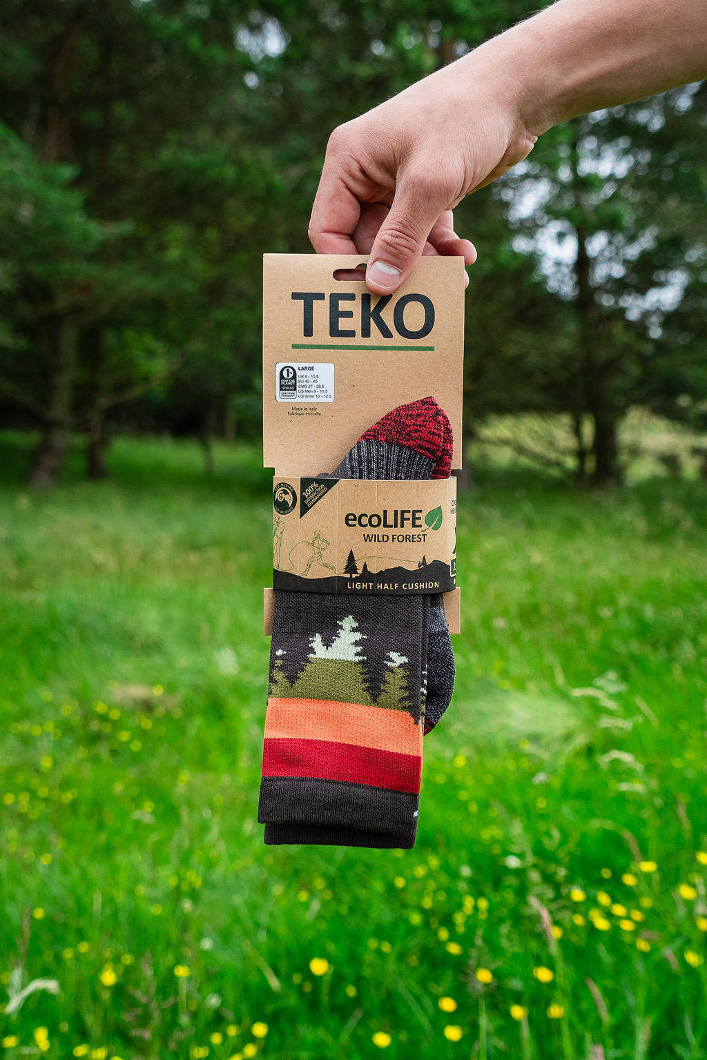 A hand holds a package of TEKO ecoLIFE wild forest knit socks against a blurred green outdoor background of trees and grass.