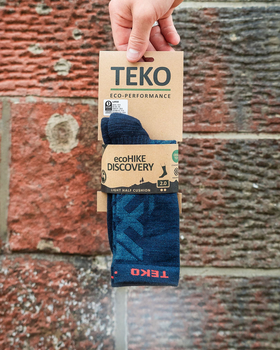 A hand holds a package of TEKO ecoHIKE DISCOVERY sock against a red brick wall background. The package contains dark blue socks with a subtle pattern.
