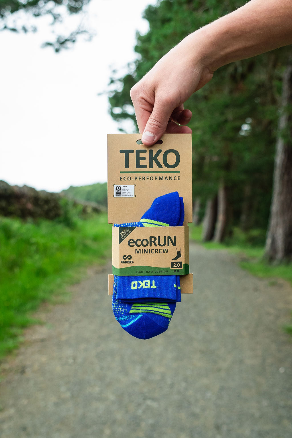 A hand holds a package of TEKO ecoRUN MiniCrew socks against a blurred outdoor background of a gravel path and green trees. The socks are blue with lime green accents, and the packaging is brown kraft paper.