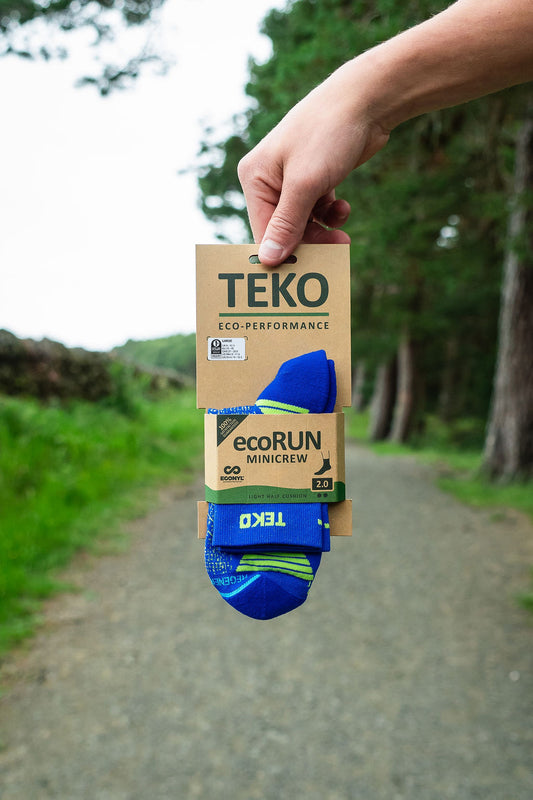 A hand holds a package of TEKO ecoRUN MiniCrew socks against a blurred outdoor background of a gravel path and green trees. The socks are blue with lime green accents, and the packaging is brown kraft paper.