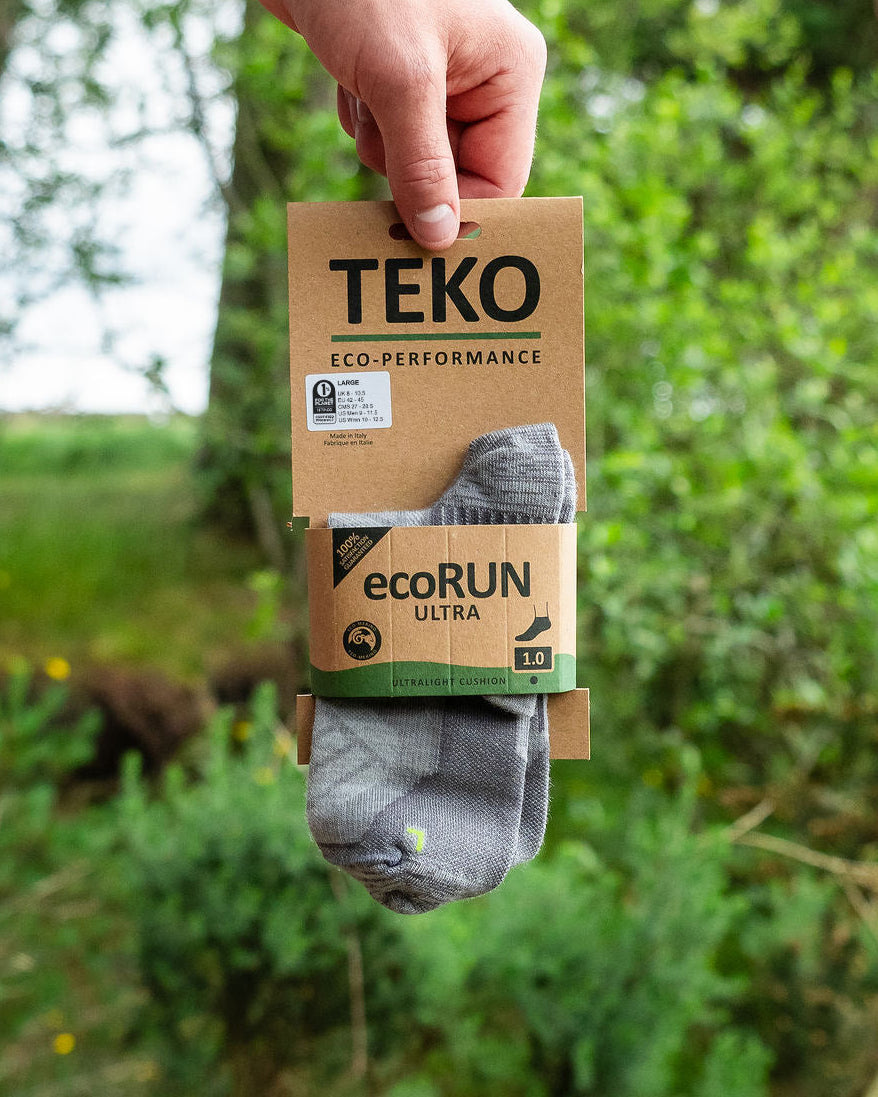 A hand holds a package of TEKO ecoRUN ultra socks against a blurred green outdoor background. The socks are gray with a small green and yellow accent, and the packaging is made of brown cardboard.