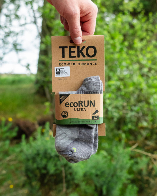 A hand holds a package of TEKO ecoRUN ultra socks against a blurred green outdoor background. The socks are gray with a small green and yellow accent, and the packaging is made of brown cardboard.
