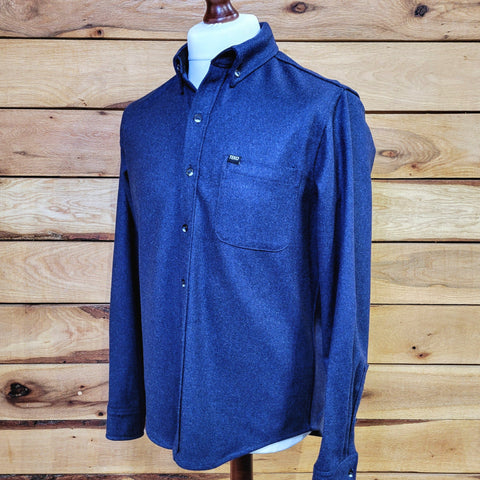 A storm blue, long-sleeved men's shirt displayed on a mannequin against a wooden backdrop, featuring a button-down collar and a chest pocket.